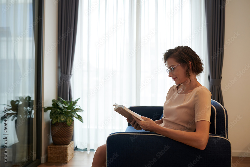 Positive middle-aged woman reading book at home when resting on sofa Positive middle-aged woman reading book at home when resting on sofa