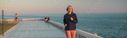 Leinwand Poster Asian sportswoman wearing black hoodie jogging on the embankment against the bac