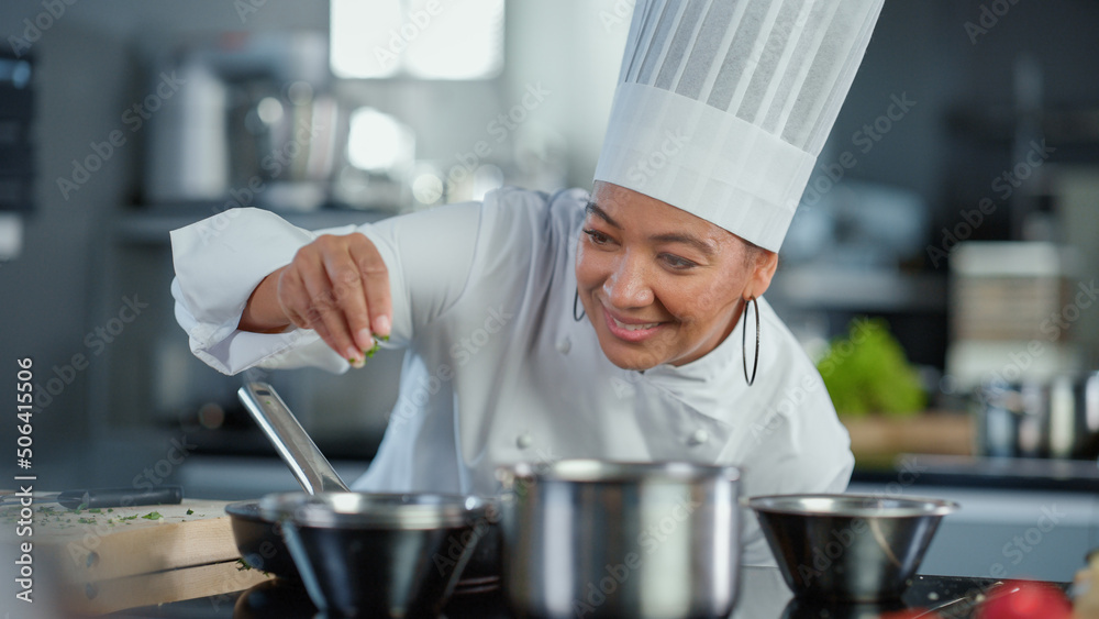 Restaurant Kitchen: Black Female Chef Fries Uses Pan, Seasons Dish with Herbs and Spices, Smiles. Professional Cooking Delicious and Traditional Authentic Food. Healthy Dishes Prepare. Medium Shot Restaurant Kitchen: Black Female Chef Fries Uses Pan, Seasons Dish with Herbs and Spices, Smiles. Professional Cooking Delicious and Traditional Authentic Food. Healthy Dishes Prepare. Medium Shot