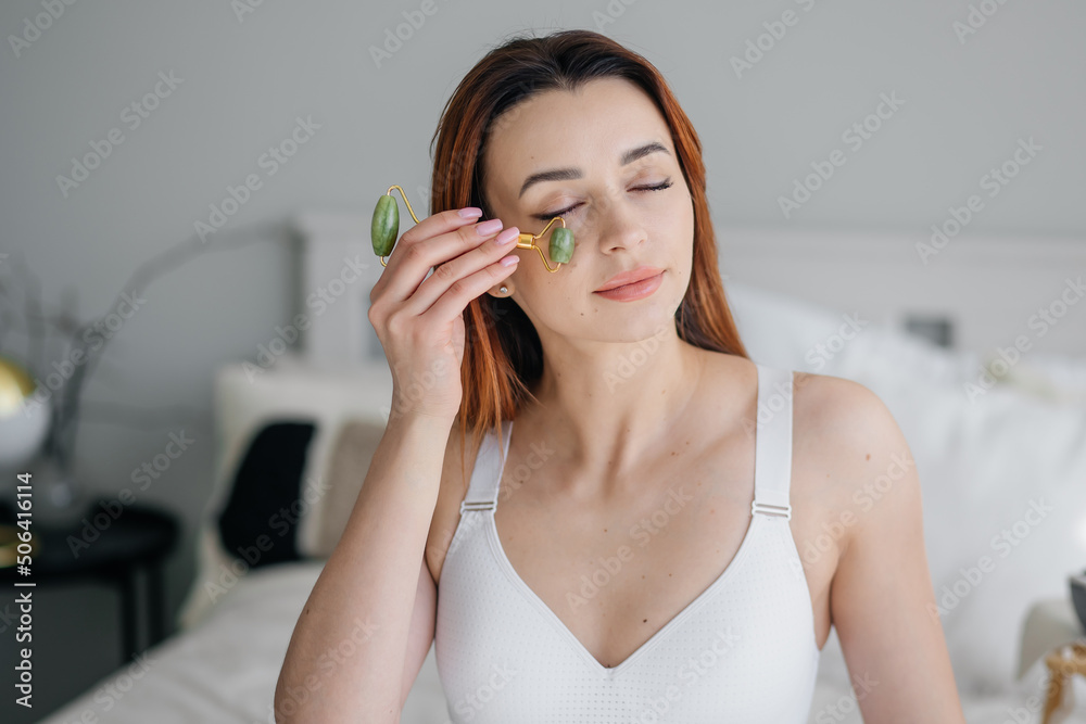 Young woman of European model appearance with blue eyes makes a facial jade massage with a roller. Bedroom with stylish interior. Young woman of European model appearance with blue eyes makes a facial jade massage with a roller. Bedroom with stylish interior.