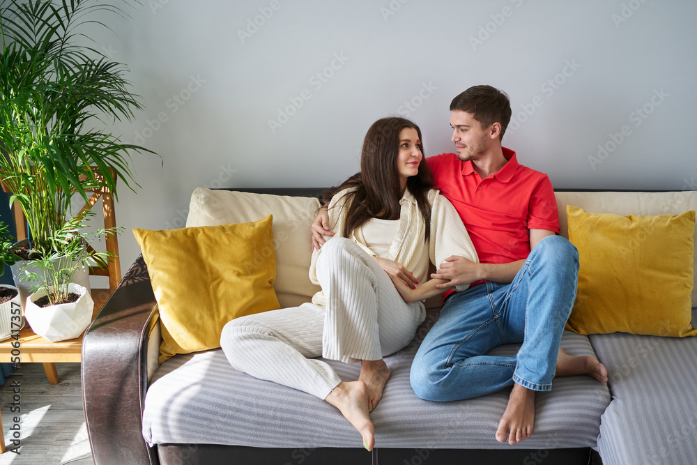 young beautiful couple sits at home on the couch and looks at each other with tenderness young beautiful couple sits at home on the couch and looks at each other with tenderness