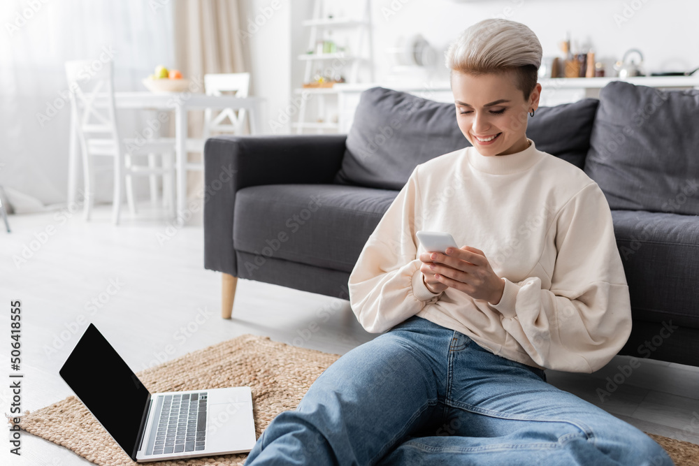 happy woman sitting on floor with mobile phone near laptop with blank screen
