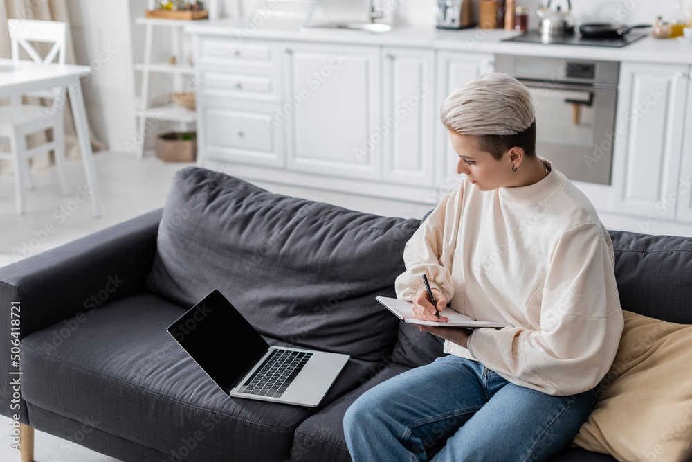 woman with trendy hairstyle writing in notebook near laptop on sofa at home