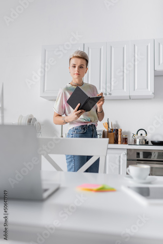 stylish woman standing with notebook in kitchen and looking at blurred laptop