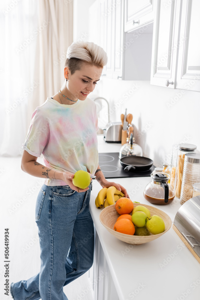 trendy young woman holding apple and bananas near bowl with fresh fruits in kitchen