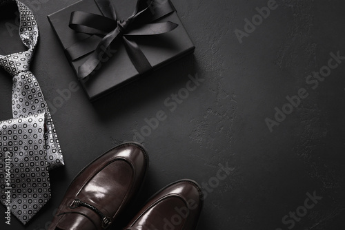 Happy Fathers Day greeting card with gift box. Set of classic mens clothes - brown shoes, tie and gift on black background. Men's accessories set. Top view. Copy space.
