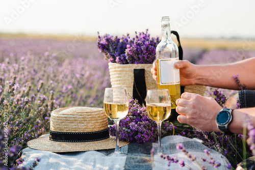 Male hands pouring white wine from a bottle into glasses on a background of a lavender field. Straw hat and basket with flowers lavender on a blanket on picnic table. Romantic evening time for dinner.