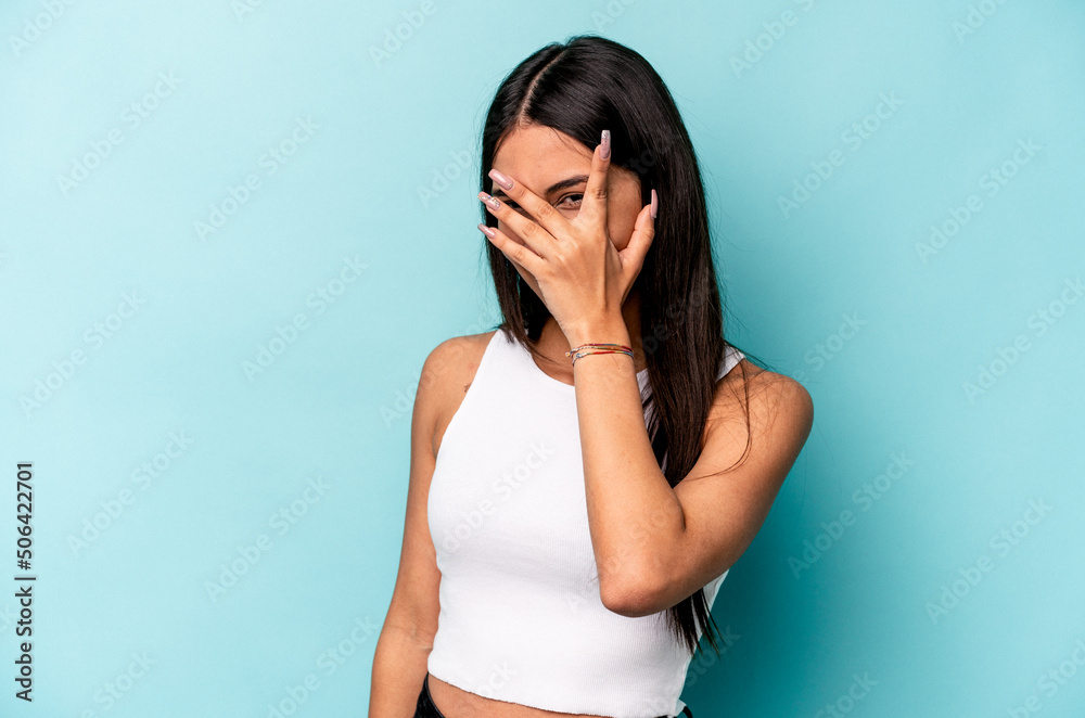 Young hispanic woman isolated on blue background blink at the camera through fingers, embarrassed covering face.