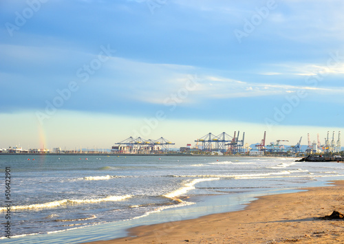 Coastline beach near seaport of Valencia. Empty beach and no people. Container crane in ship port for shipping containers. Sea with storm waves near dock. Ports and Terminal, Maritime Industry.