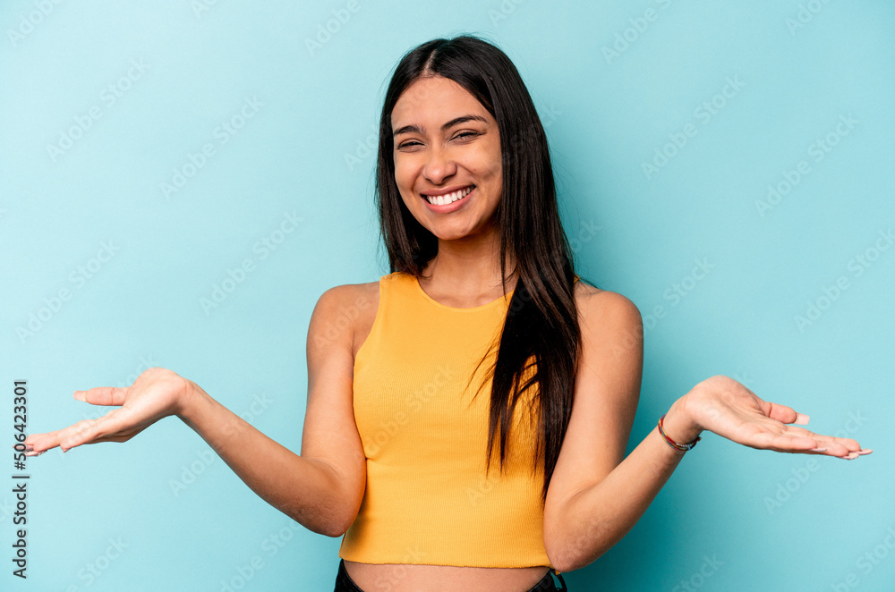 Young hispanic woman isolated on blue background makes scale with arms, feels happy and confident. Young hispanic woman isolated on blue background makes scale with arms, feels happy and confident.