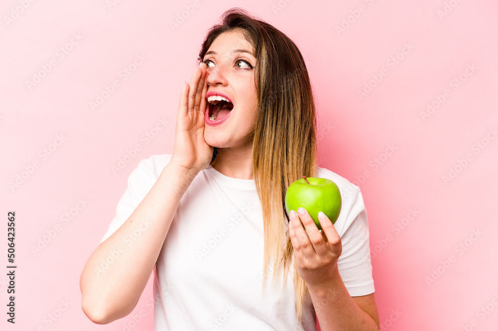 Young caucasian woman holding an apple isolated on pink background shouting and holding palm near opened mouth. Young caucasian woman holding an apple isolated on pink background shouting and holding palm near opened mouth.