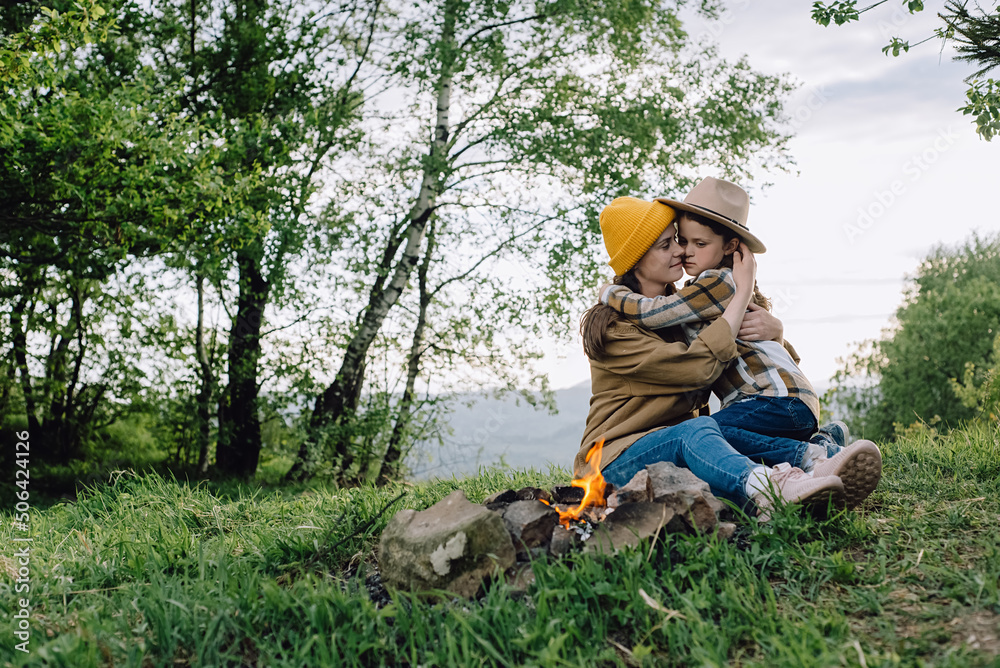 Happy loving young mom and cute little daughter child in hat sit near bonfire on background green forest. Smiling small kid hugs mother, enjoy summer nature. Camping holiday with kids in summer nature Happy loving young mom and cute little daughter child in hat sit near bonfire on background green forest. Smiling small kid hugs mother, enjoy summer nature. Camping holiday with kids in summer nature