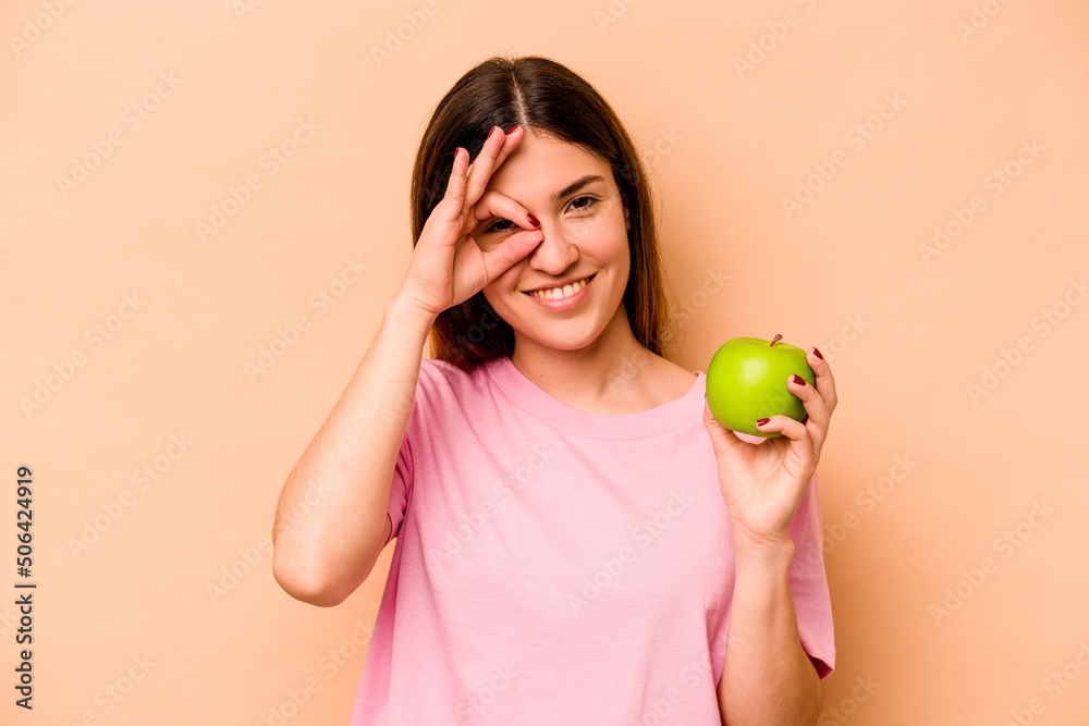 Young hispanic woman holding an apple isolated on beige background excited keeping ok gesture on eye. Young hispanic woman holding an apple isolated on beige background excited keeping ok gesture on eye.