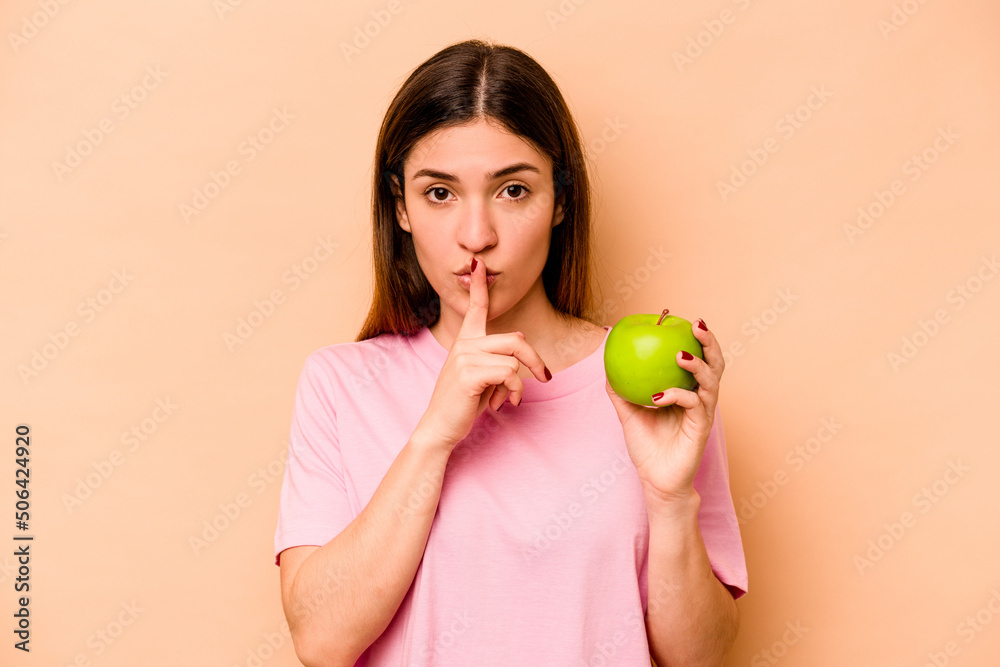 Young hispanic woman holding an apple isolated on beige background keeping a secret or asking for silence. Young hispanic woman holding an apple isolated on beige background keeping a secret or asking for silence.