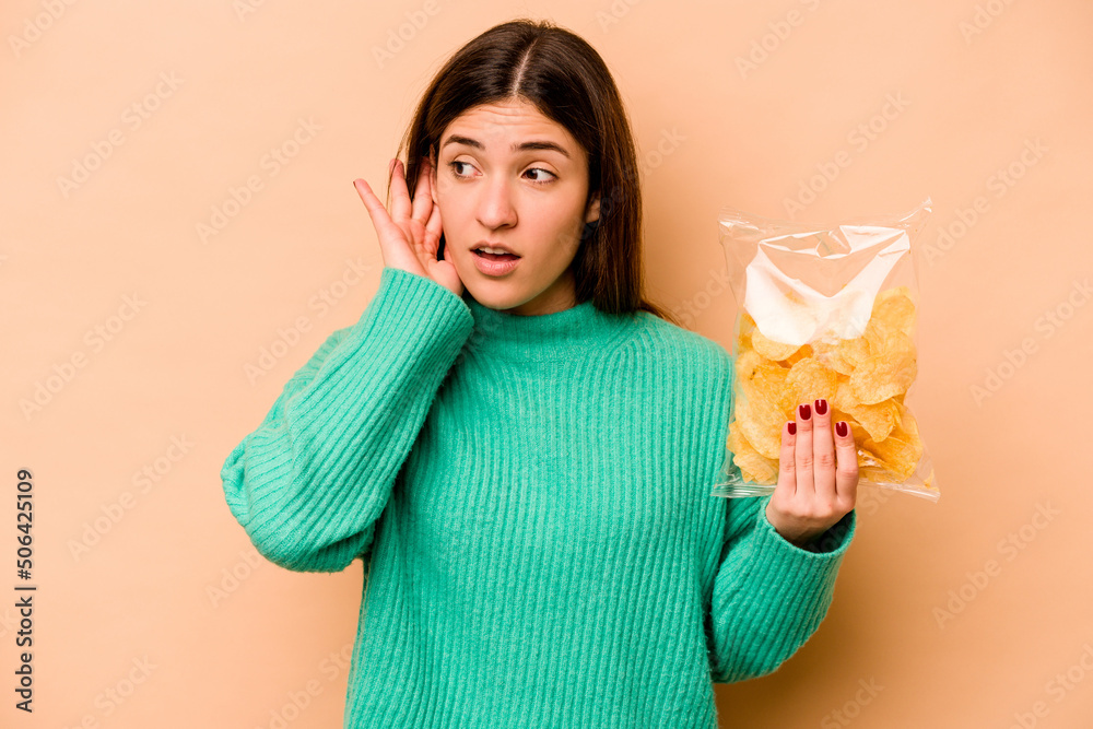 Young hispanic woman holding a bag of chips isolated on beige background trying to listening a gossip. Young hispanic woman holding a bag of chips isolated on beige background trying to listening a gossip.