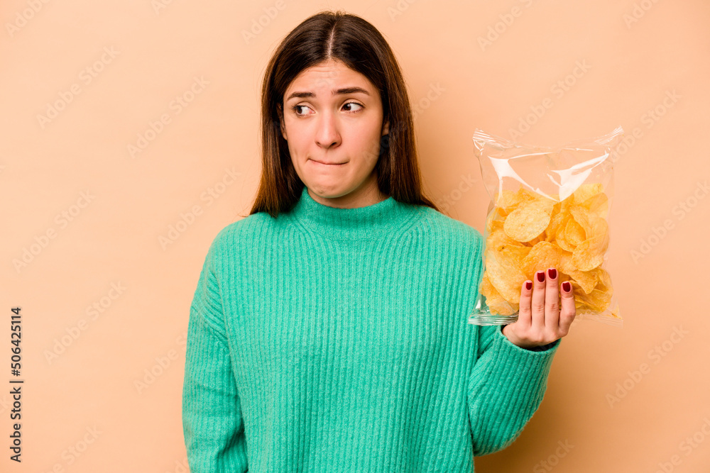 Young hispanic woman holding a bag of chips isolated on beige background confused, feels doubtful and unsure. Young hispanic woman holding a bag of chips isolated on beige background confused, feels doubtful and unsure.