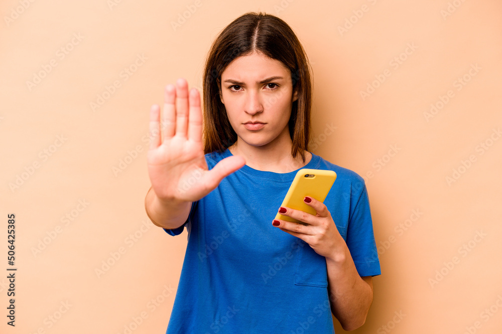 Young caucasian woman holding mobile phone isolated on beige background standing with outstretched hand showing stop sign, preventing you.