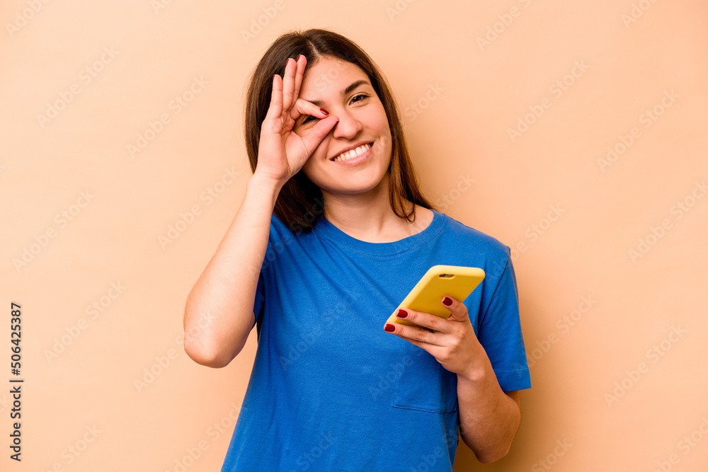 Young caucasian woman holding mobile phone isolated on beige background excited keeping ok gesture on eye. Young caucasian woman holding mobile phone isolated on beige background excited keeping ok gesture on eye.