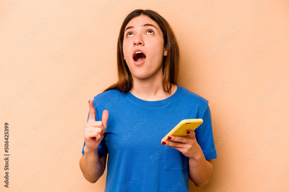 Young caucasian woman holding mobile phone isolated on beige background pointing upside with opened mouth. Young caucasian woman holding mobile phone isolated on beige background pointing upside with opened mouth.