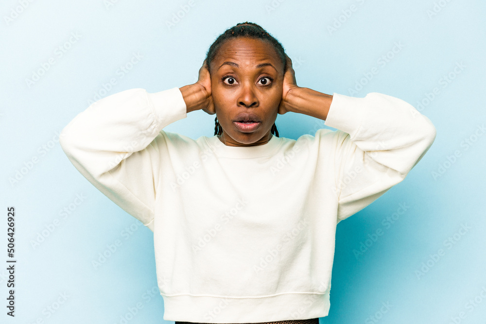 Young african american woman isolated on blue background covering ears with hands trying not to hear too loud sound.
