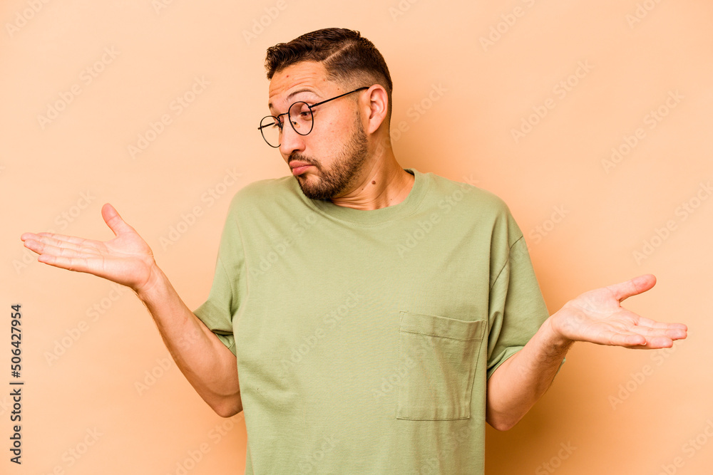 Young hispanic man isolated on beige background confused and doubtful shrugging shoulders to hold a copy space.