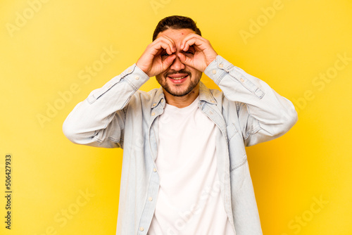 Young hispanic man isolated on yellow background showing okay sign over eyes © Asier