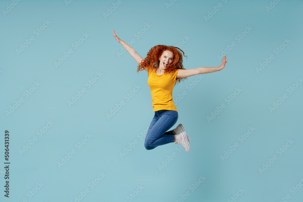 Full body young excited happy redhead woman 20s wearing yellow t-shirt jump high with outstretched arms hands isolated on plain light pastel blue background studio portrait. People lifestyle concept.