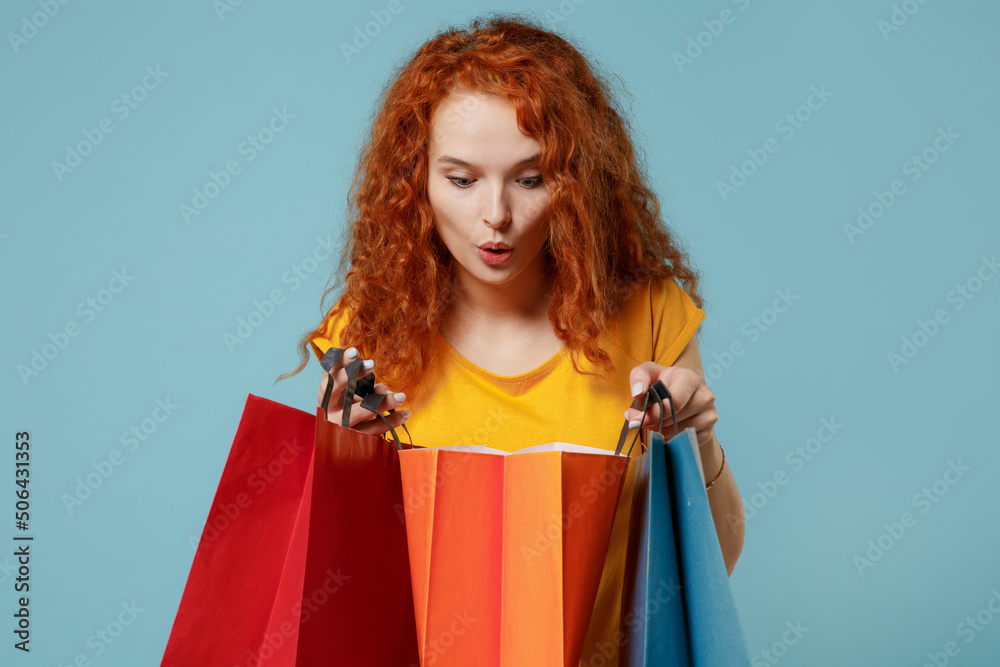 Young shocked happy redhead woman 20s wear yellow t-shirt holding package bags with purchases after shopping isolated on plain light pastel blue background studio portrait. People lifestyle concept. Young shocked happy redhead woman 20s wear yellow t-shirt holding package bags with purchases after shopping isolated on plain light pastel blue background studio portrait. People lifestyle concept.