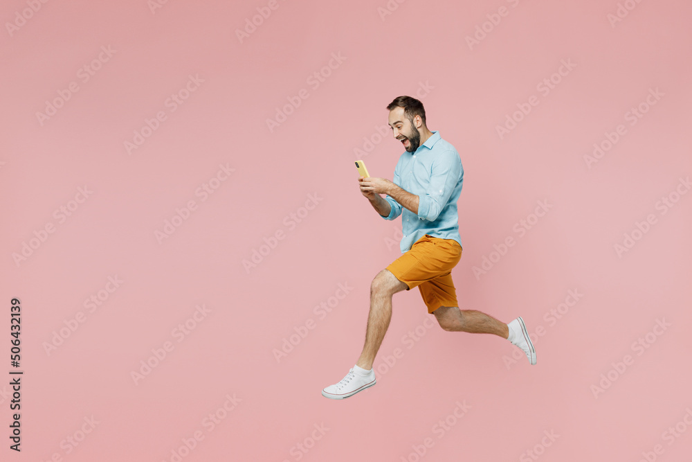 Full body side view fun young man 20s wearing classic blue shirt hold use mobile cell phone jump high run fast isolated on plain pastel light pink background studio portrait. People lifestyle concept.