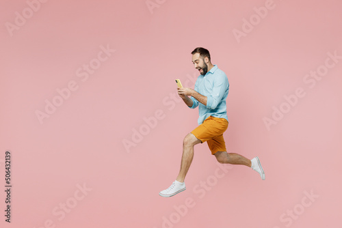 Full body side view fun young man 20s wearing classic blue shirt hold use mobile cell phone jump high run fast isolated on plain pastel light pink background studio portrait. People lifestyle concept. © ViDi Studio