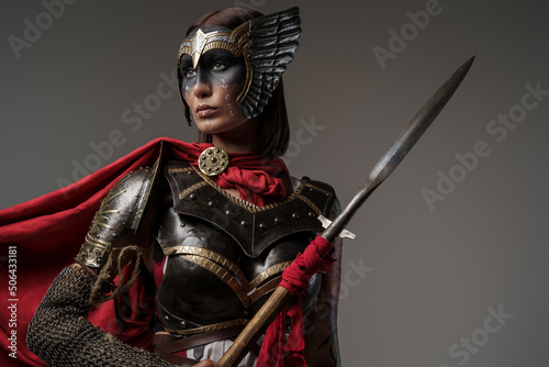 Foto Shot of female barbarian holding spear dressed in steel armor with helmet against grey background
