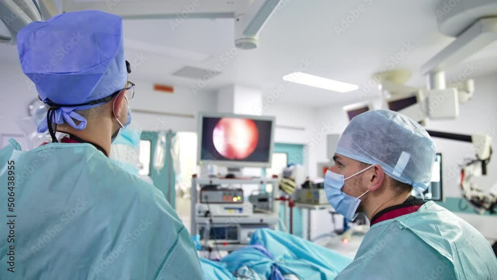 Surgeons follow the screen at operation. Doctors stand with their backs to the camera and look at the monitor. Modern surgery room at backdrop in blur. Surgeons follow the screen at operation. Doctors stand with their backs to the camera and look at the monitor. Modern surgery room at backdrop in blur.