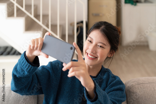 Photo of young asian woman taking selfie on cell phone while sitting on couch in bright apartment