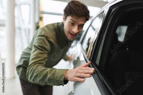 Happy young man touching new automobile of his dreams, buying modern car at dealership, selective focus