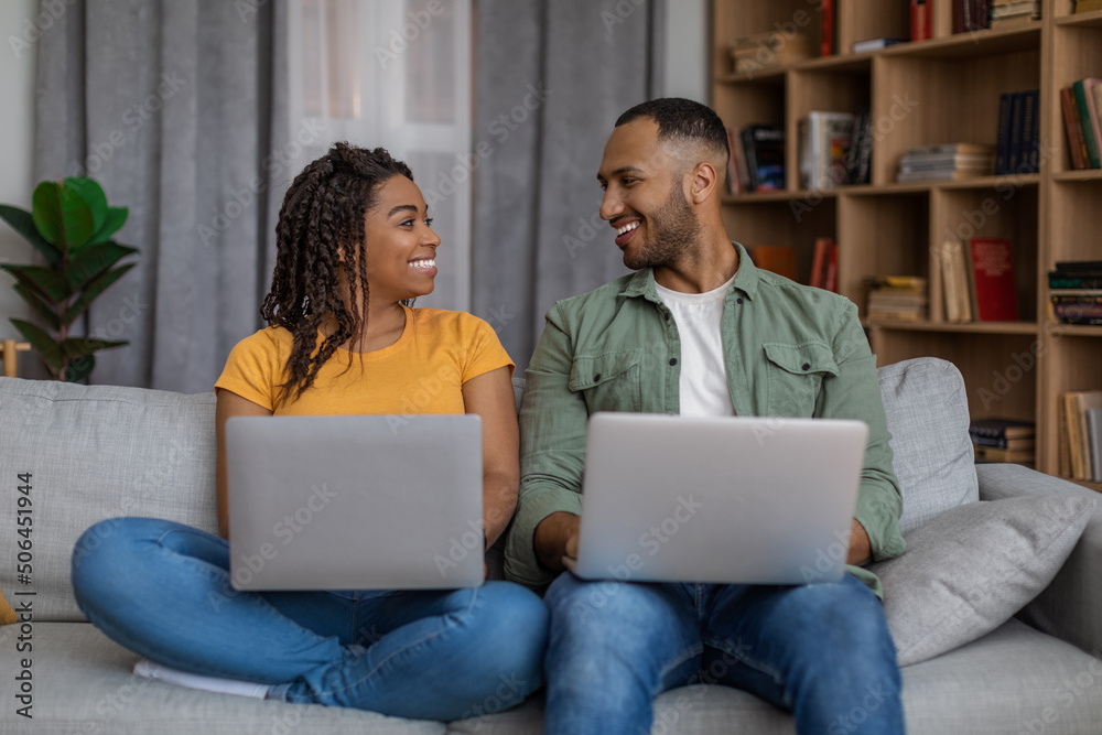 Young black couple using laptop computers, looking and smiling to each other, sitting on couch and surfing internet Young black couple using laptop computers, looking and smiling to each other, sitting on couch and surfing internet