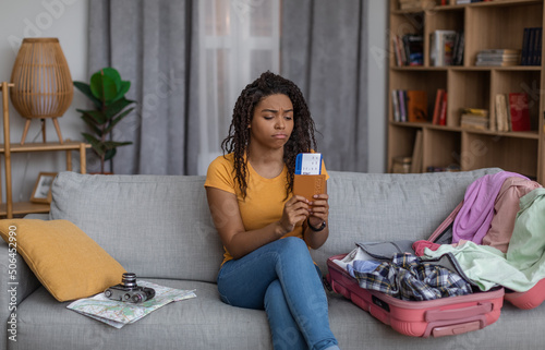 Crying black woman with travel tickets sitting on sofa with unpacked suitcase, being upset of flight cancellation photo