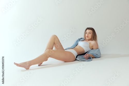 Portrait of young beautiful woman posing in white cotton underwear, lying on floor isolated on grey background