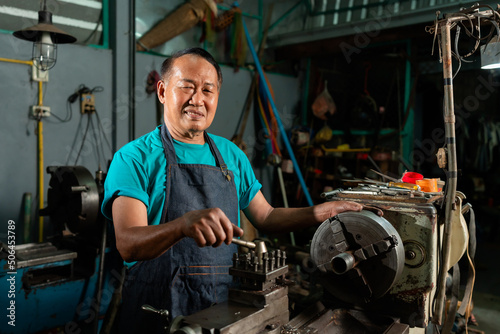 A senior Asian craftsman, standing closely and carefully controlling a lathe, inside a small factory of his family business.