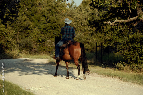 Western ranch lifestyle with cowboy horseback riding on bay mare horse through Texas summer gravel path outdoors. © ccestep8