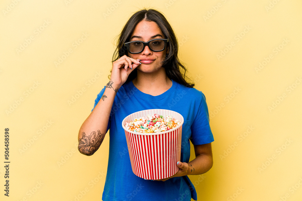 Young hispanic woman holding popcorn isolated on yellow background with fingers on lips keeping a secret.
