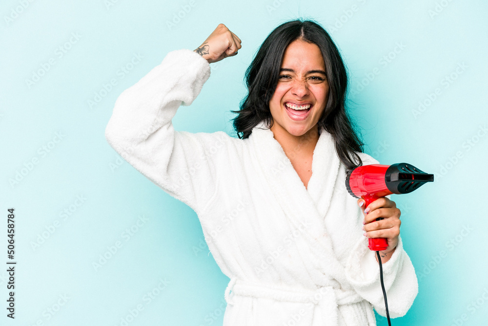 Young hispanic woman holding dryer isolated on blue background raising fist after a victory, winner concept. Young hispanic woman holding dryer isolated on blue background raising fist after a victory, winner concept.