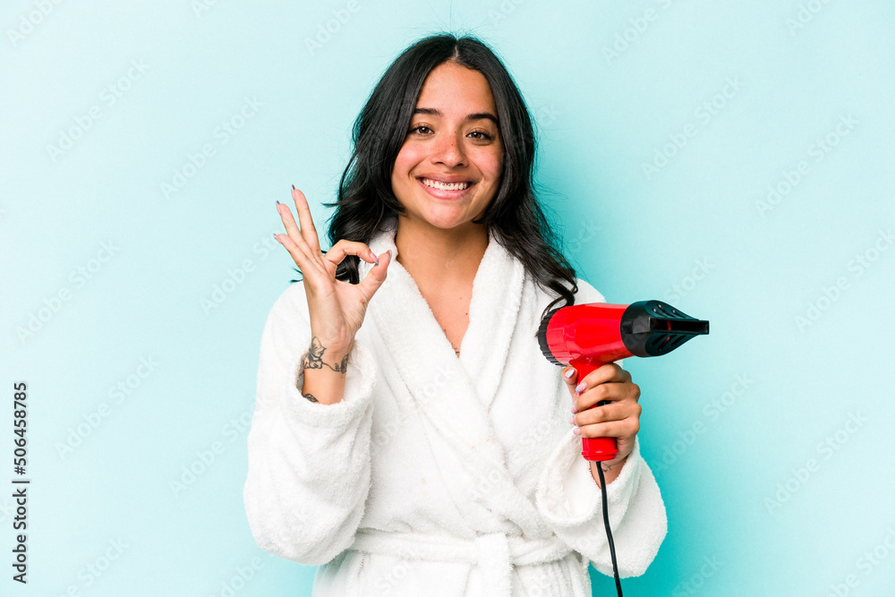 Young hispanic woman holding dryer isolated on blue background cheerful and confident showing ok gesture. Young hispanic woman holding dryer isolated on blue background cheerful and confident showing ok gesture.