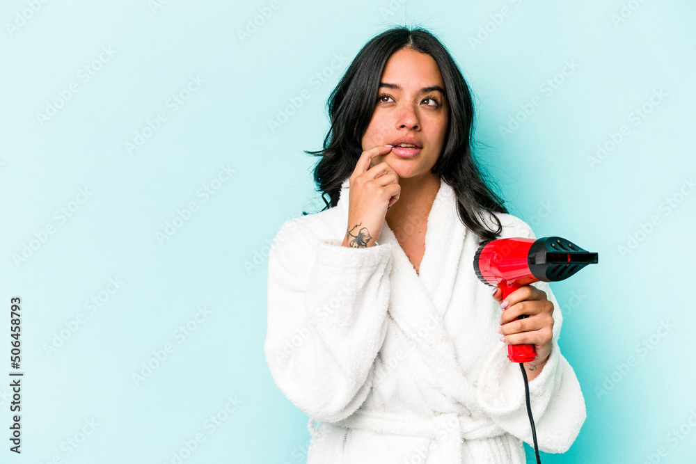 Young hispanic woman holding dryer isolated on blue background relaxed thinking about something looking at a copy space. Young hispanic woman holding dryer isolated on blue background relaxed thinking about something looking at a copy space.