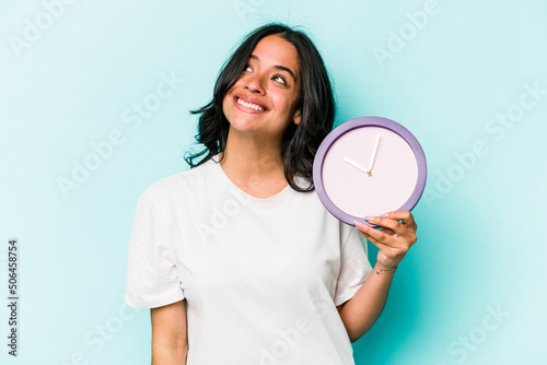 Young hispanic woman holding a clock isolated on blue background dreaming of achieving goals and purposes