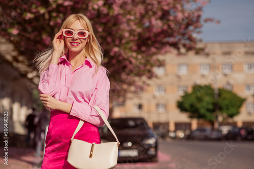 Φωτογραφία Happy smiling fashionable woman wearing trendy total pink outfit with sunglasses, shirt, satin mini skirt, holding white faux leather bag, posing in street of city