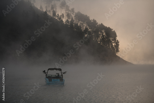 rear view of motorboat floating on the water against the backdrop of silhouette of hill in fog
