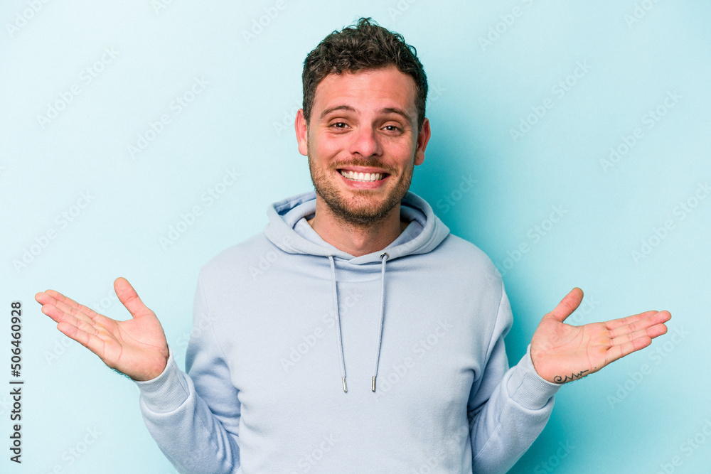 Young caucasian man isolated on blue background makes scale with arms, feels happy and confident.