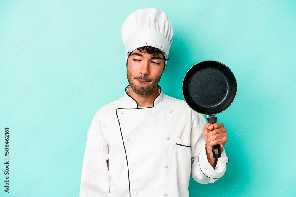 Young caucasian chef man holding flying pan isolated on blue background confused, feels doubtful and unsure. Young caucasian chef man holding flying pan isolated on blue background confused, feels doubtful and unsure.