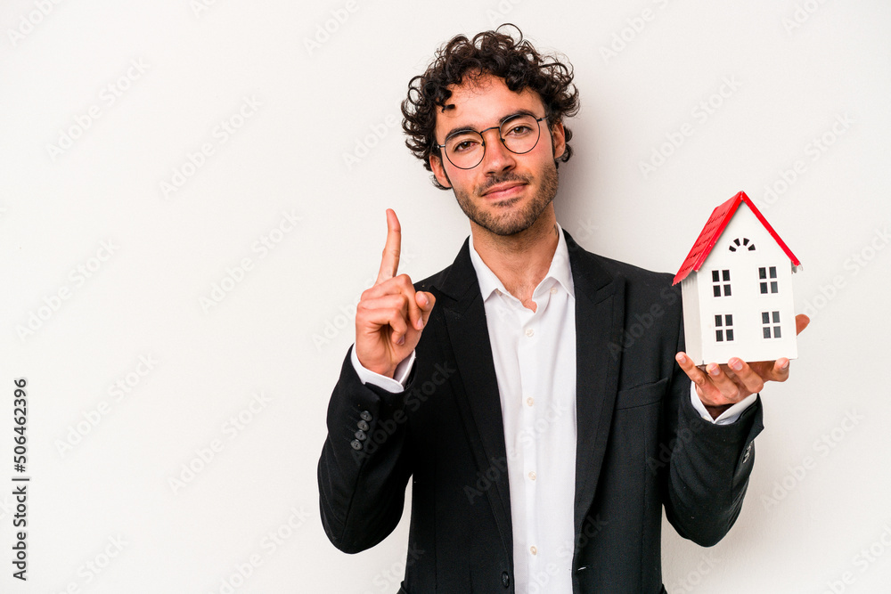 Young caucasian business man holding a toy house isolated on white background showing number one with finger. Young caucasian business man holding a toy house isolated on white background showing number one with finger.