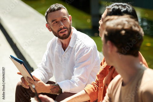 Young businessman in white shirt holding tablet computer when talking to colleagues outdoors